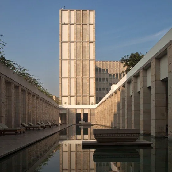 Modern building with a tall central tower, surrounding courtyard with a water feature, lounge chairs, and building reflections in the water.