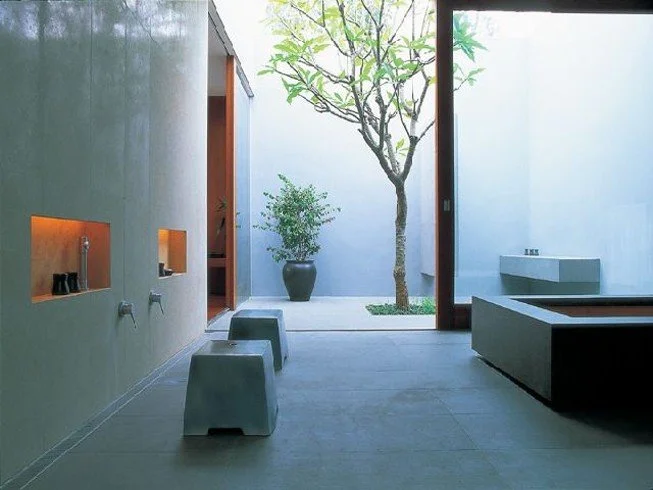 Minimalist bathroom with gray walls, a window view of a tree, a small plant in a black pot, two gray stools, a white rectangular bathtub, and a modern sink.
