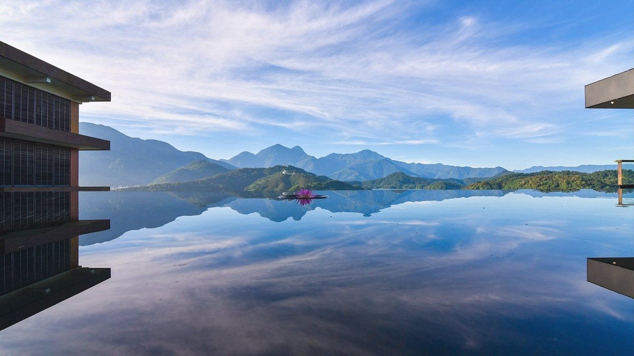 View of blue sky, mountains, and greenery reflected in the still water of an infinity pool.