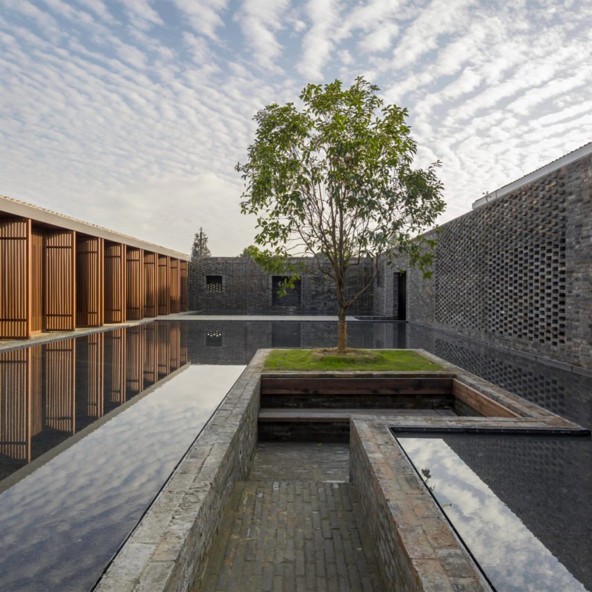 A modern courtyard with a central tree, surrounded by brick walls, a reflecting pool, and a clear sky with scattered clouds.