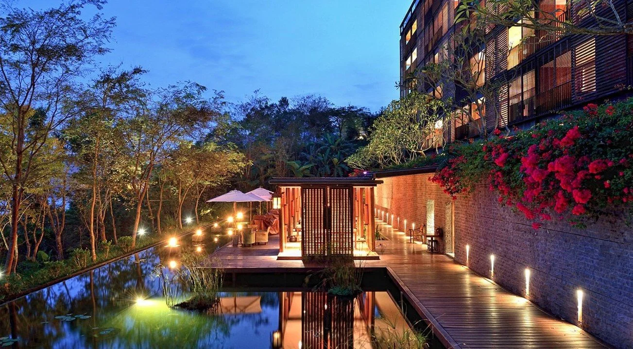 A modern outdoor patio area at dusk with a reflective pool, wooden pathway, illuminated seating, lush trees, and a multi-story building with a balcony and flowering plants.