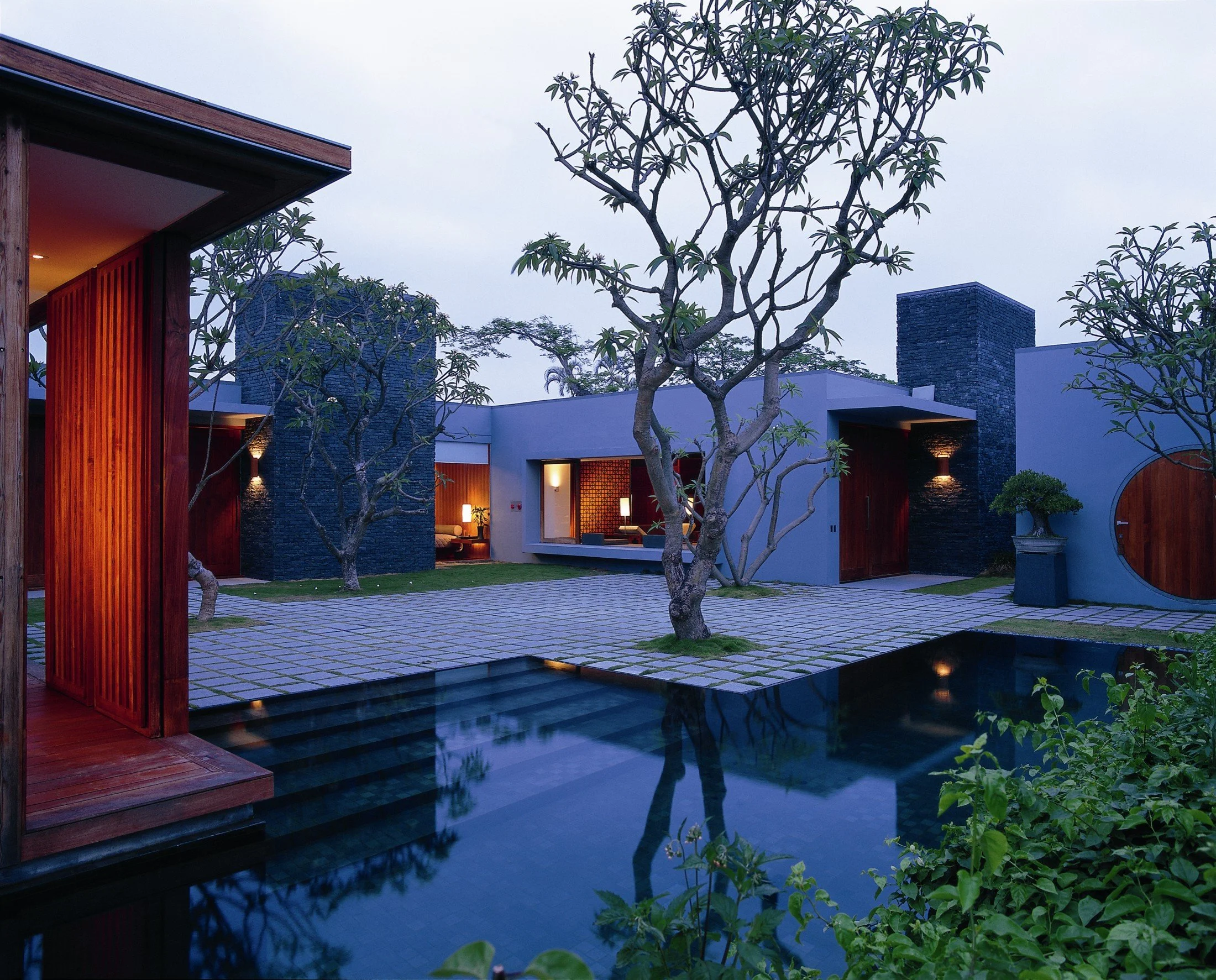 Modern house with a courtyard featuring a swimming pool, trees, outdoor lighting, and a mix of wooden and stone architectural elements during evening.