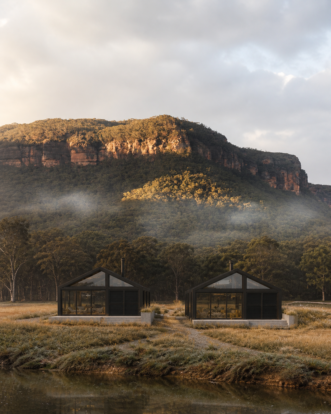 Two modern black glass houses with outdoor steps are situated in a grassy field near a small water body. In the background, there is a forested mountain with cloud and mist at the base, under a cloudy sky.