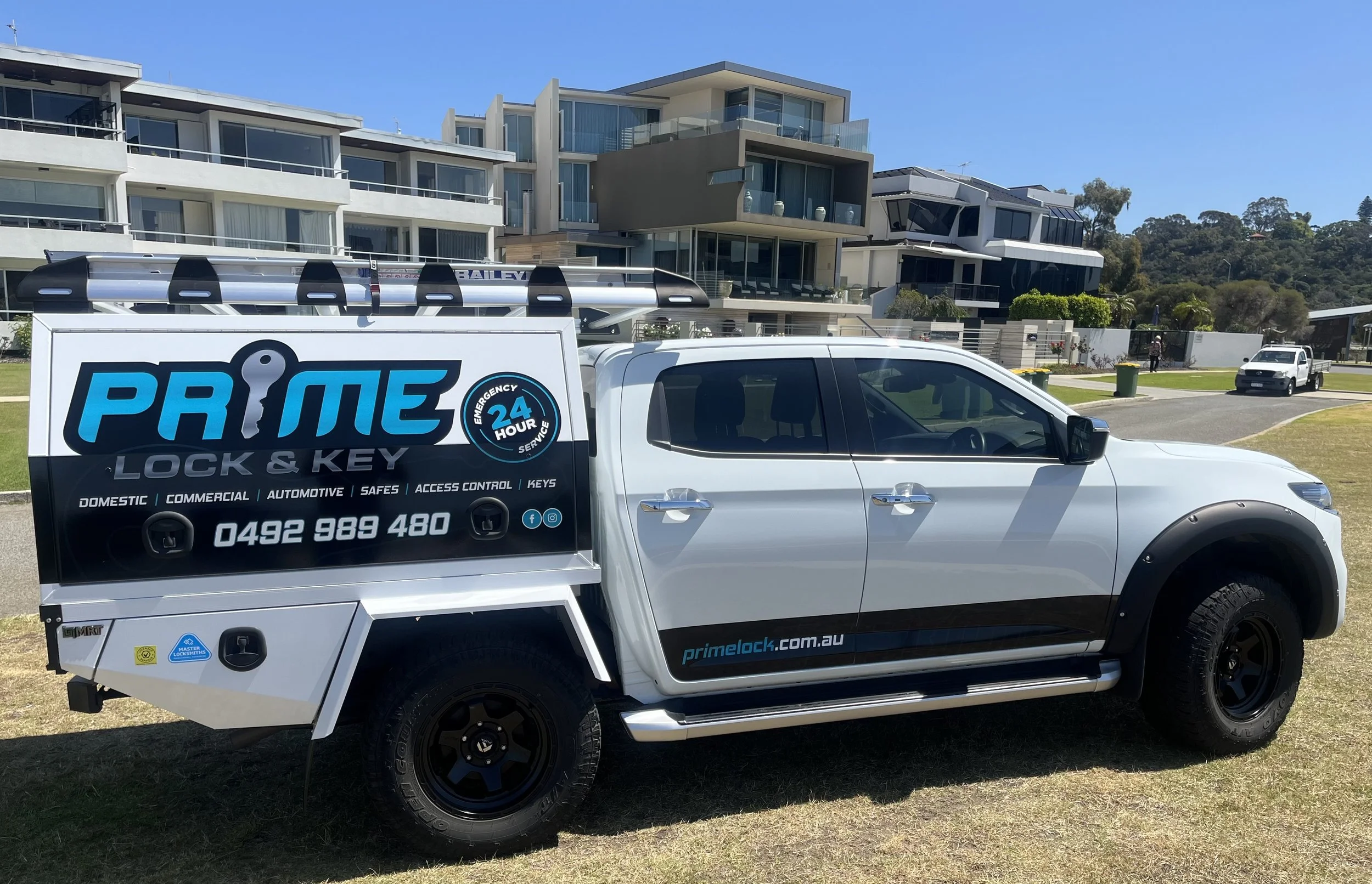 A white service truck parked on a grassy area in front of modern residential buildings. The truck's side displays the logo for Prime Lock & Key, a 24-hour emergency locksmith service, with contact details and service offerings.