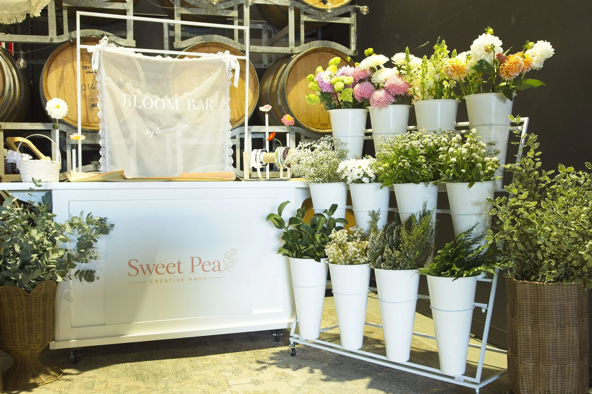 Display stand with various white and pink flowers in white pots, a sign with 'BLOOM BAR by Sweet Pea', and wooden barrels in the background.