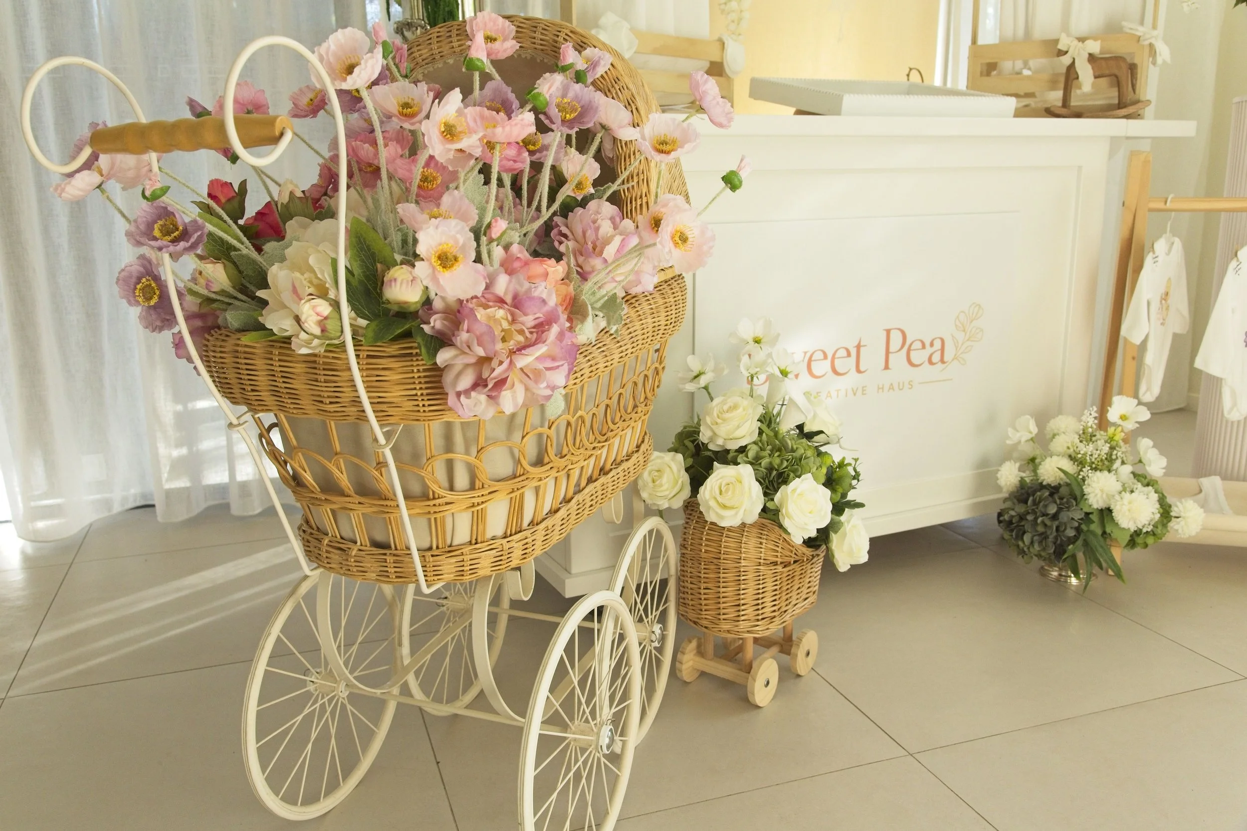 A decorative display with a vintage white metal doll stroller filled with pink and cream artificial flowers, accompanied by wicker baskets with flowers on a tiled floor at a floral shop or boutique, with a white counter and clothing rack in the background.