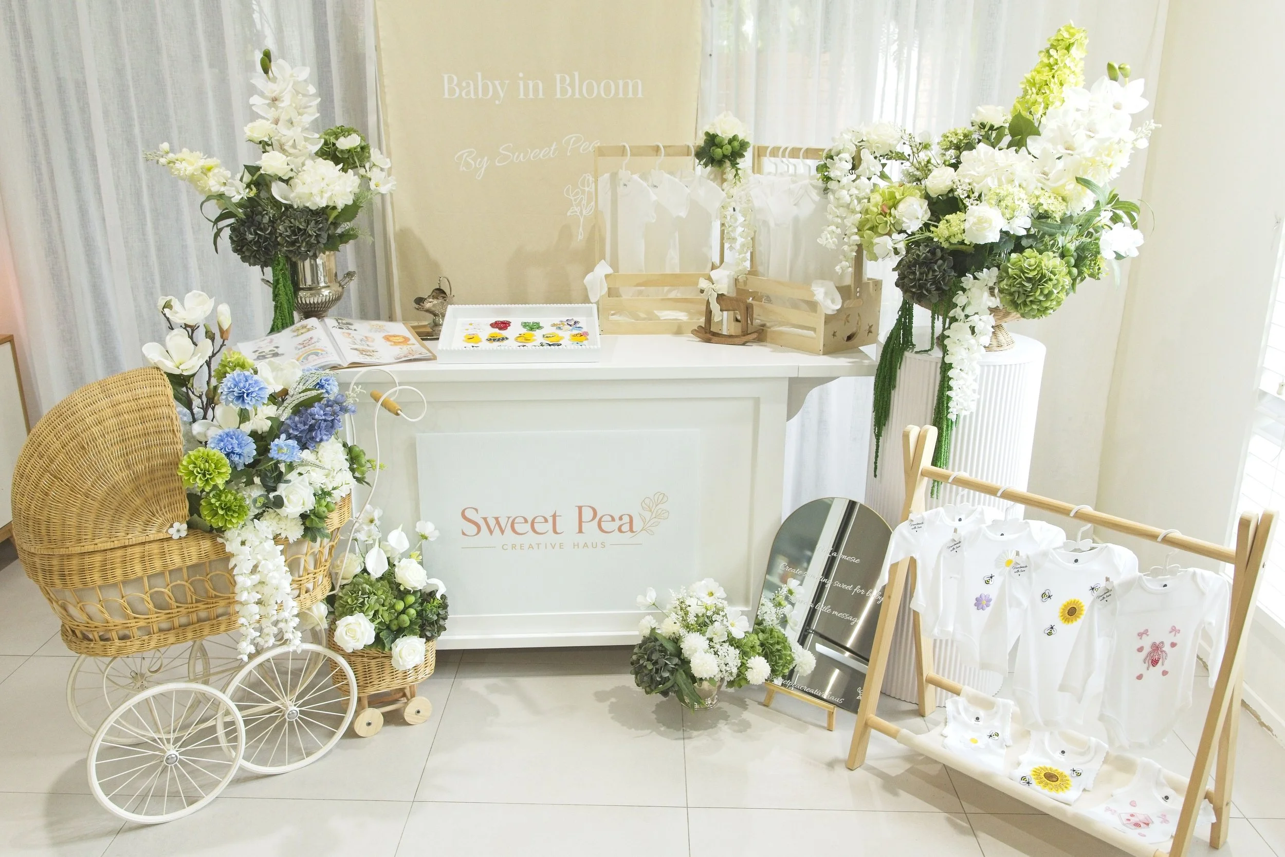 Display of floral arrangements, a miniature vintage white stroller filled with white flowers, a table with baby clothes, and a sign reading 'Baby in Bloom' at a baby shower or celebration event.