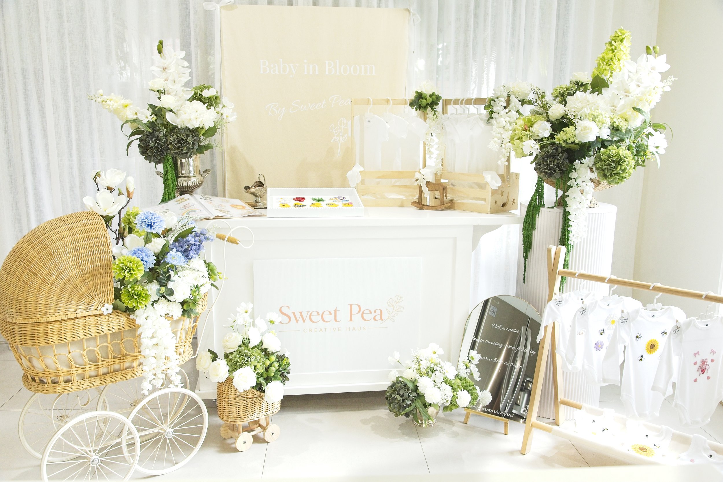 Display of white floral arrangements and decorations, including a wicker cart, floral baskets, and flower-adorned clothing, at a softly lit event space with a sign reading 'Baby in Bloom' by Sweet Pea.