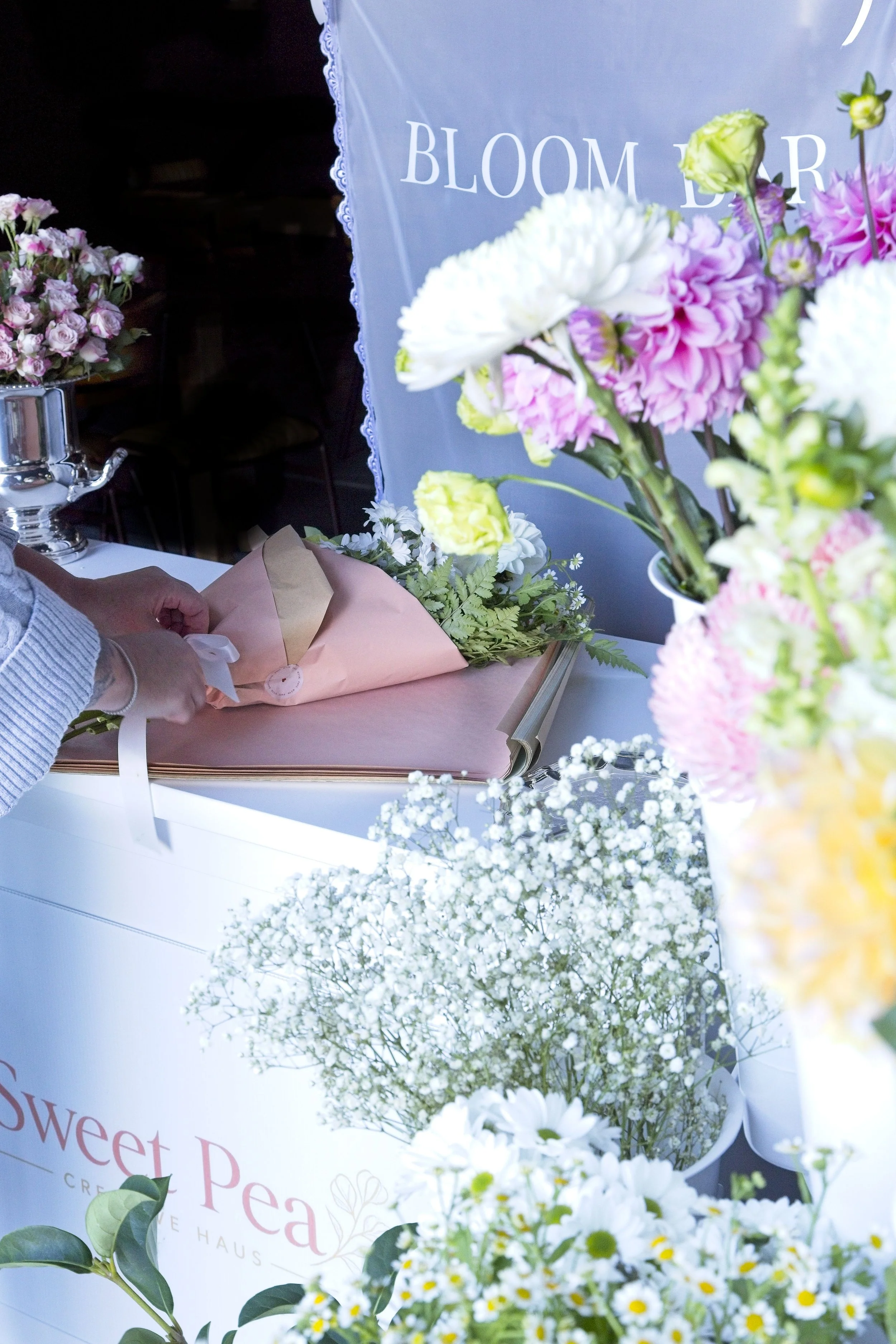 Various colorful flower arrangements, including pink and white flowers, on display at a flower shop or event with a partially visible sign reading "BLOOM" and