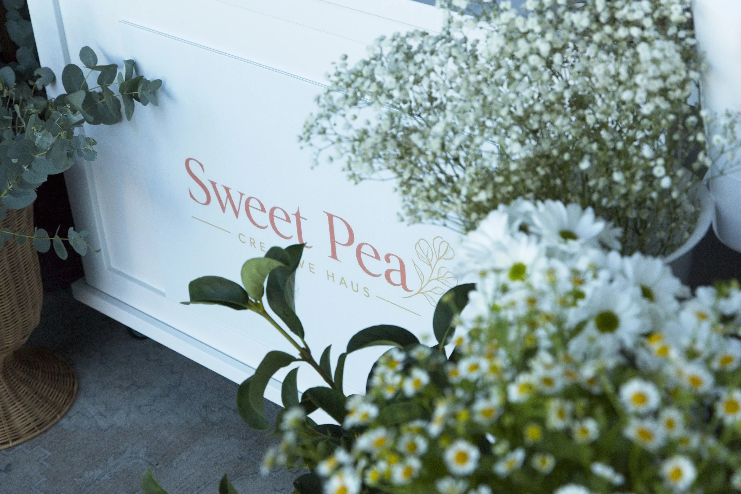 A floral display with a white sign reading "Sweet Pea" and various white and green flowers and leaves.