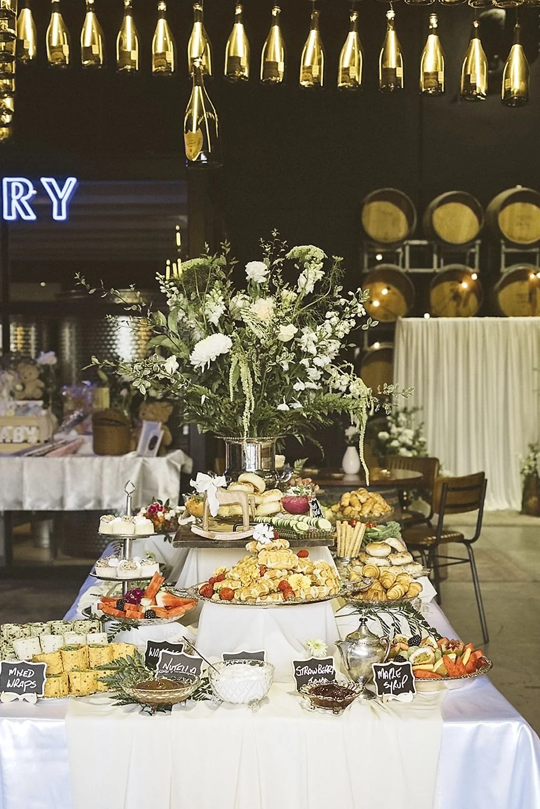 Dessert and snack table with a large floral arrangement at a celebration event with champagne bottles overhead.