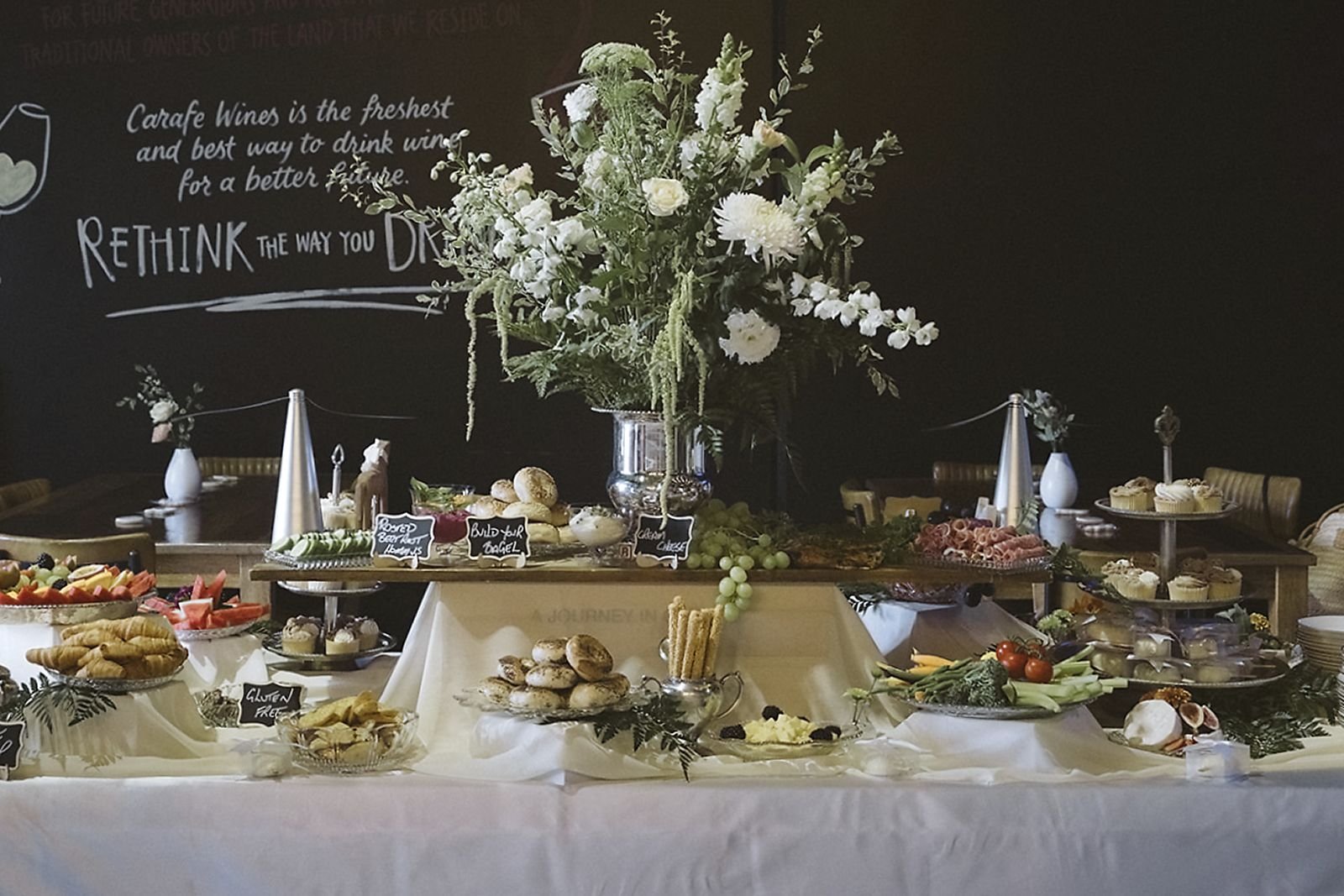 Buffet table with a large floral centerpiece in a silver vase, surrounded by various dishes including fruits, bread, cheese, and desserts, with a chalkboard wall in the background.