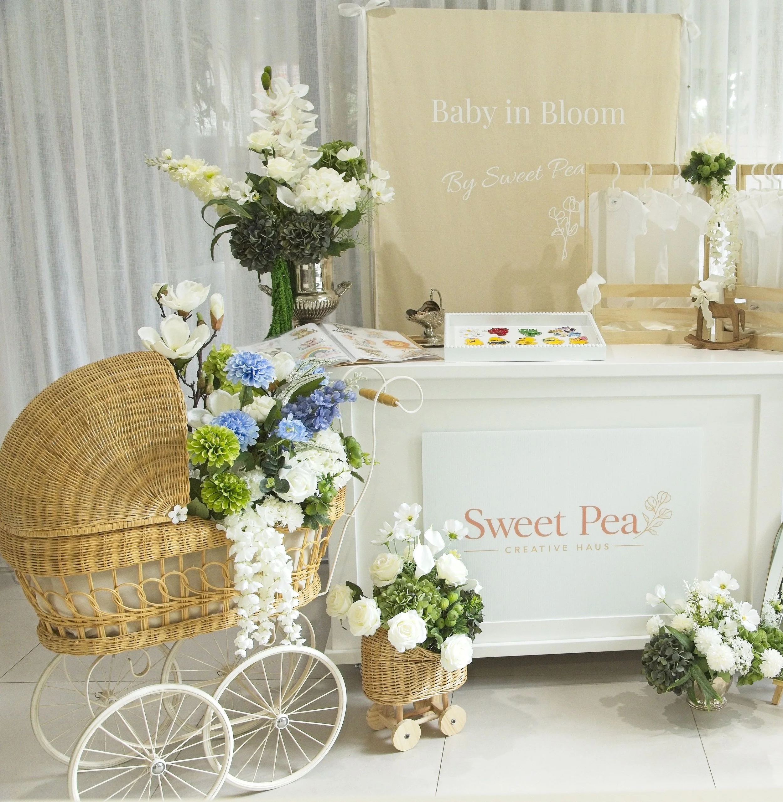 Decorative display featuring white flowers, a wicker cart, and a sign that reads "Baby in Bloom by Sweet Pea" with additional floral arrangements around.