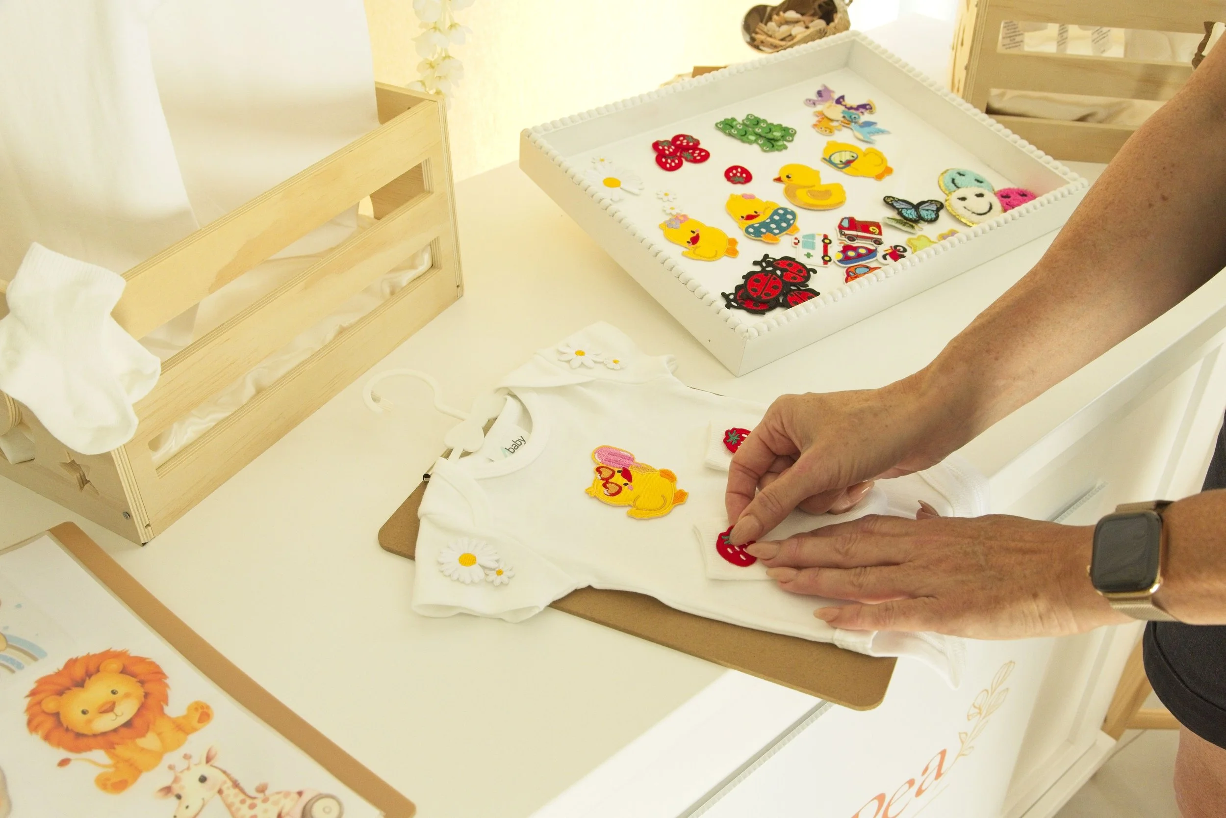 Person arranging colorful embroidered patches, including animals and flowers, on a white baby onesie at a craft station with wicker containers and creative decorations.