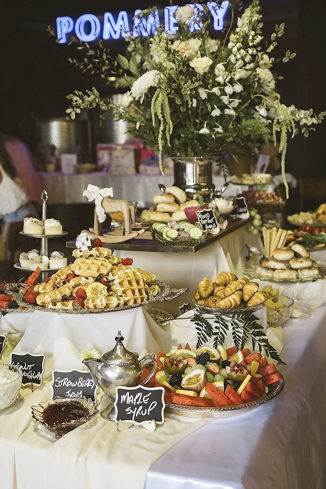 A dessert table with various pastries, fruits, and snacks, decorated with a large flower arrangement, and a glowing blue sign that says 'POMEKY'.