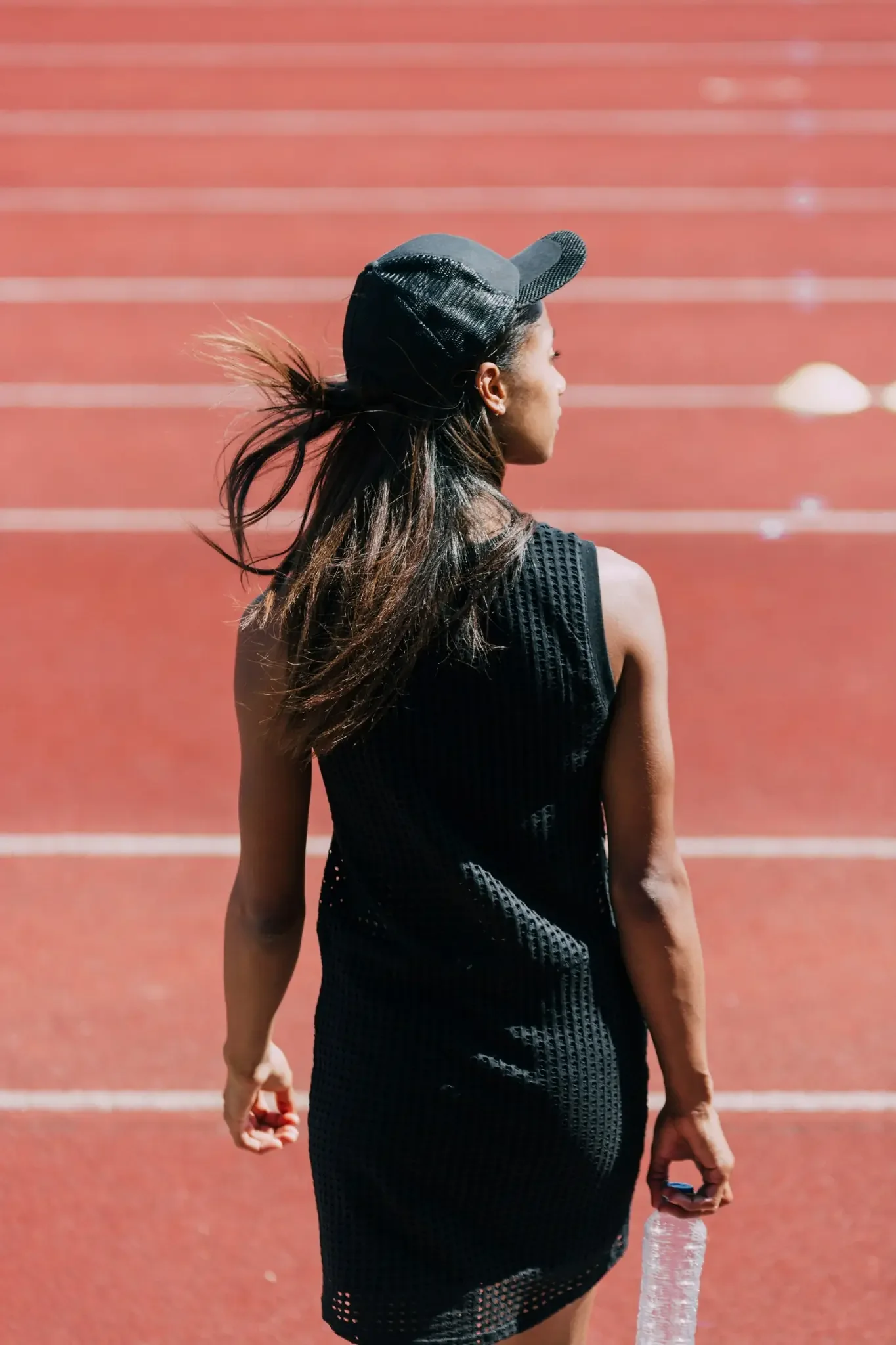 Woman in black athletic wear and a cap walking on a red running track while holding a water bottle after training.