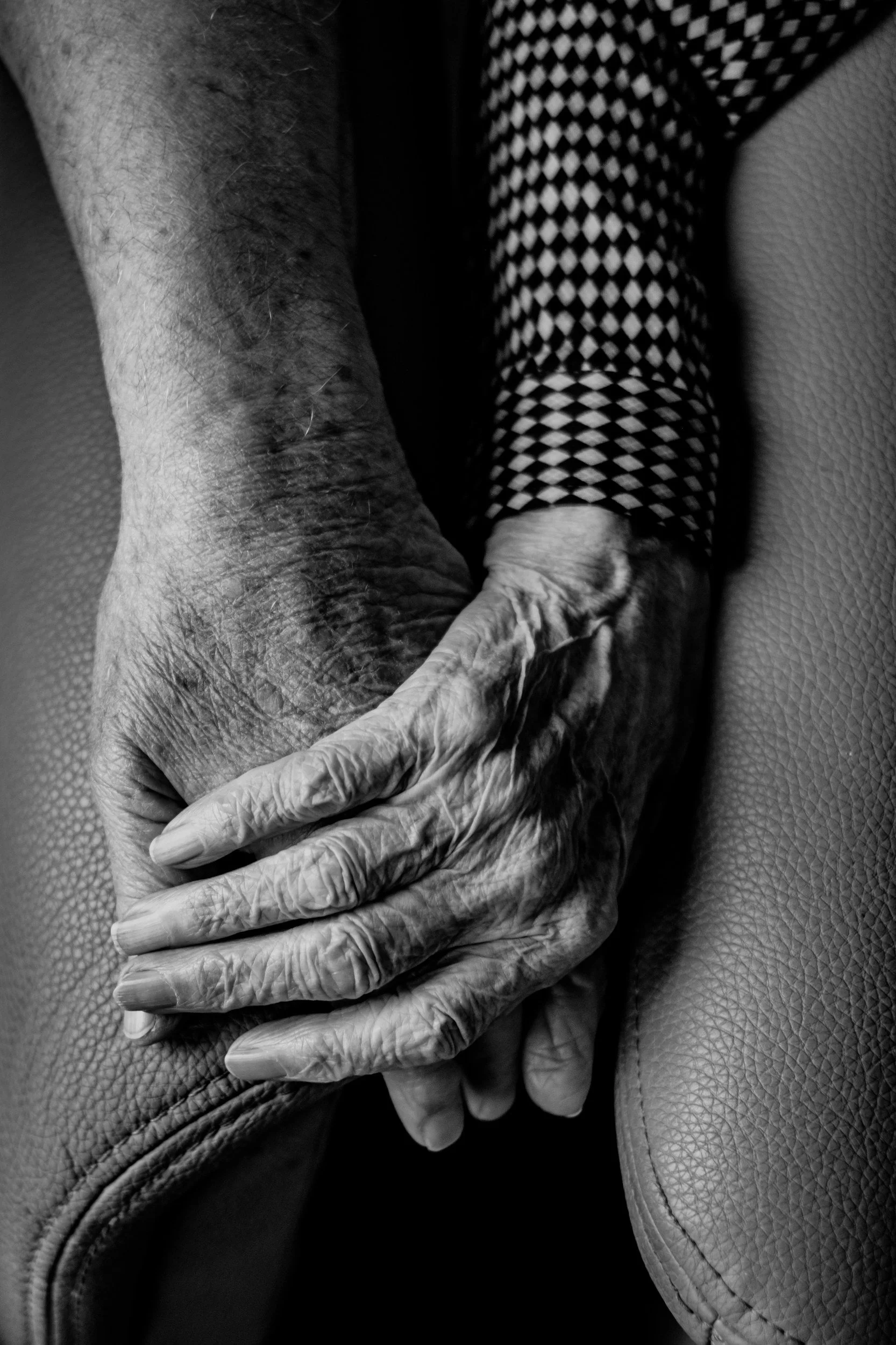 Close-up black and white photograph of two elderly hands clasped together, one hand resting on top of the other, with a textured leather surface seen beneath them.