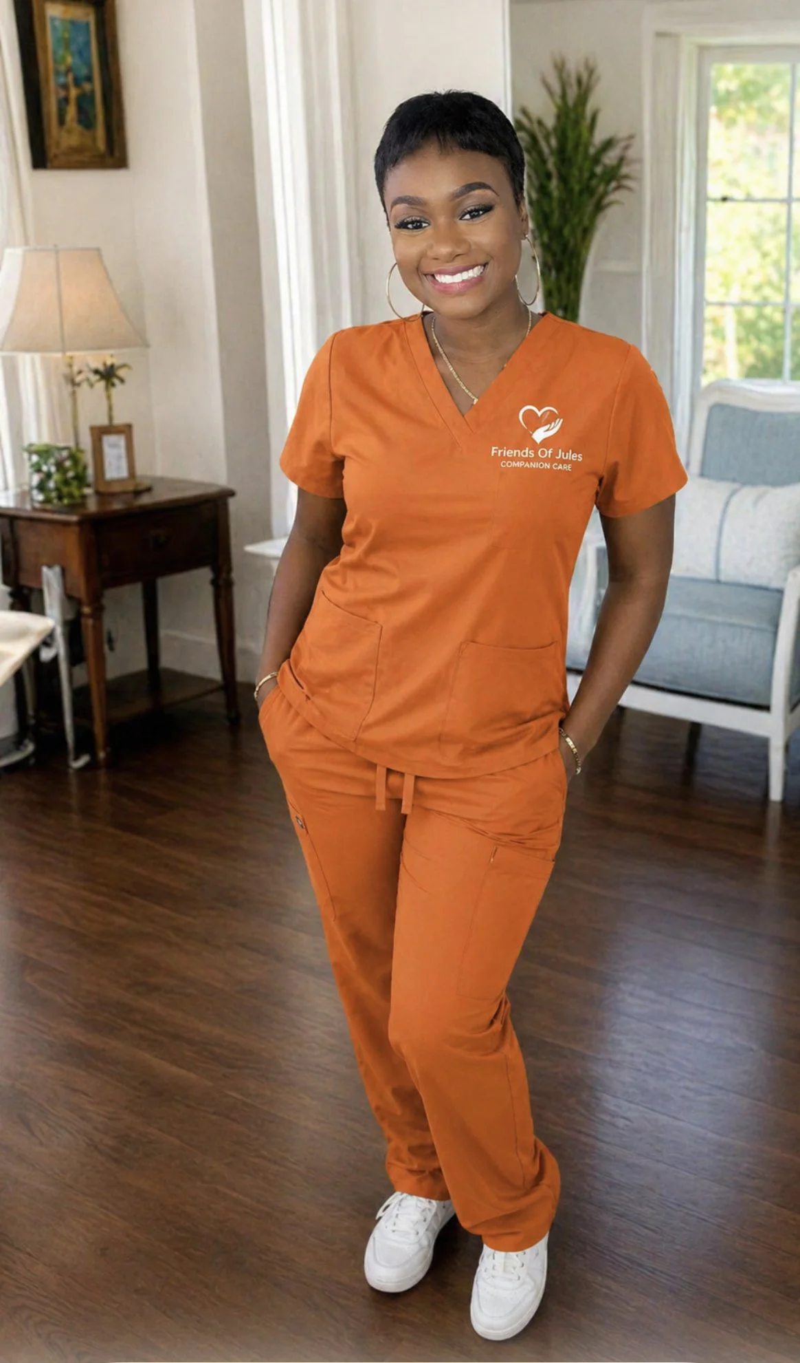 A woman in orange scrubs with a logo that reads 'Friends Of Jules Companion Care' standing in a living room with wood flooring, a side table, a lamp, a large window, and a green plant in the background.