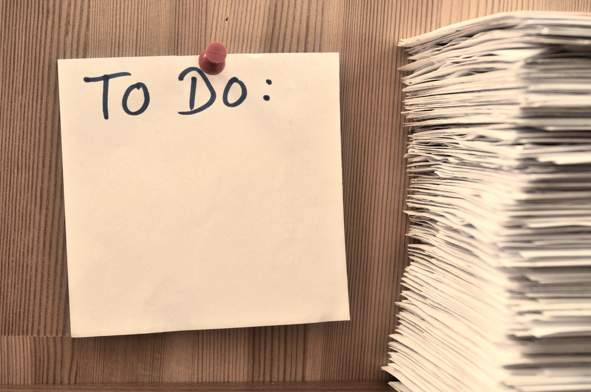 A wooden desk with a white note paper pinned with a red magnet that reads 'TO DO:' in handwritten black marker, next to a large stack of papers.