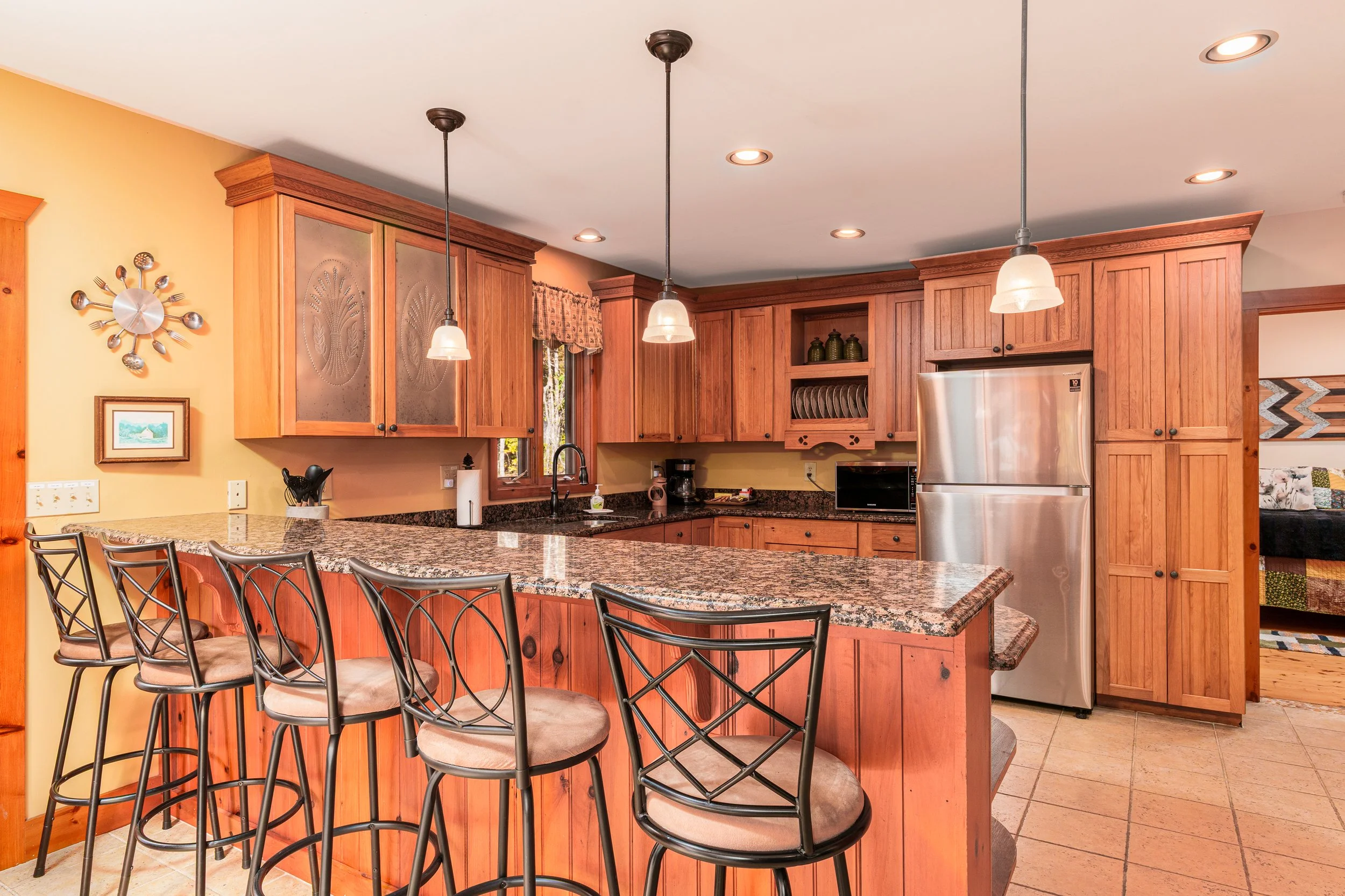A modern kitchen with wooden cabinets, a granite countertop island with several chairs, stainless steel refrigerator, pendant lights, a window with a valance, and wooden flooring.