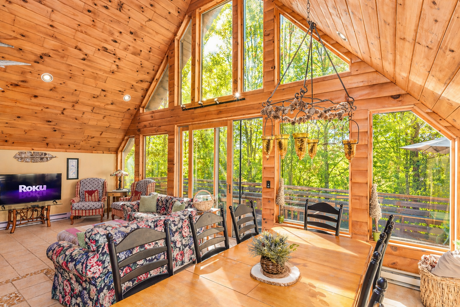 Interior view of a sunlit wooden living and dining room with large windows showing green trees outside, featuring a wooden dining table with a floral centerpiece, black chairs, a colorful floral sofa, a TV, and armchairs.
