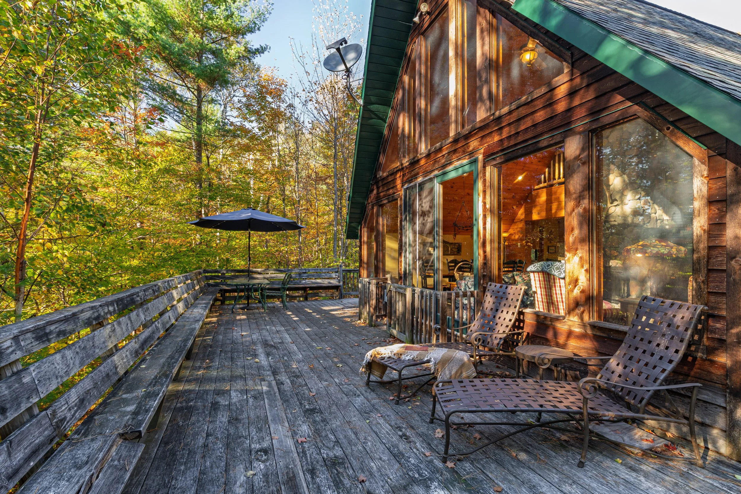 A rustic wooden Airbnb with large glass windows and a deck, surrounded by trees with autumn foliage. There is a patio table with a black umbrella and several chairs on the deck.