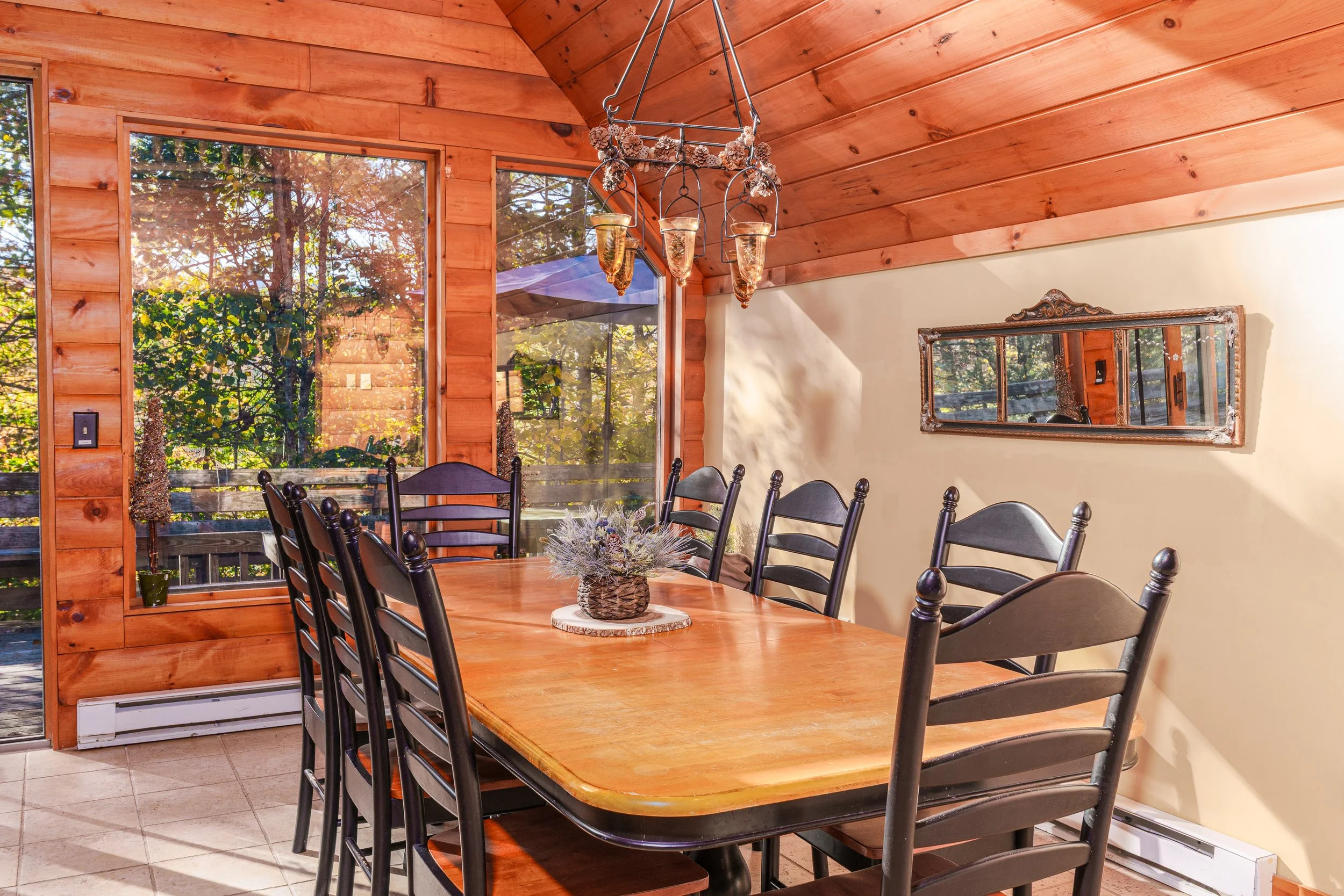 A wooden dining room table with black chairs, a flower arrangement, large windows showing trees outside, a pinecone chandelier, and a mirror on the wall.