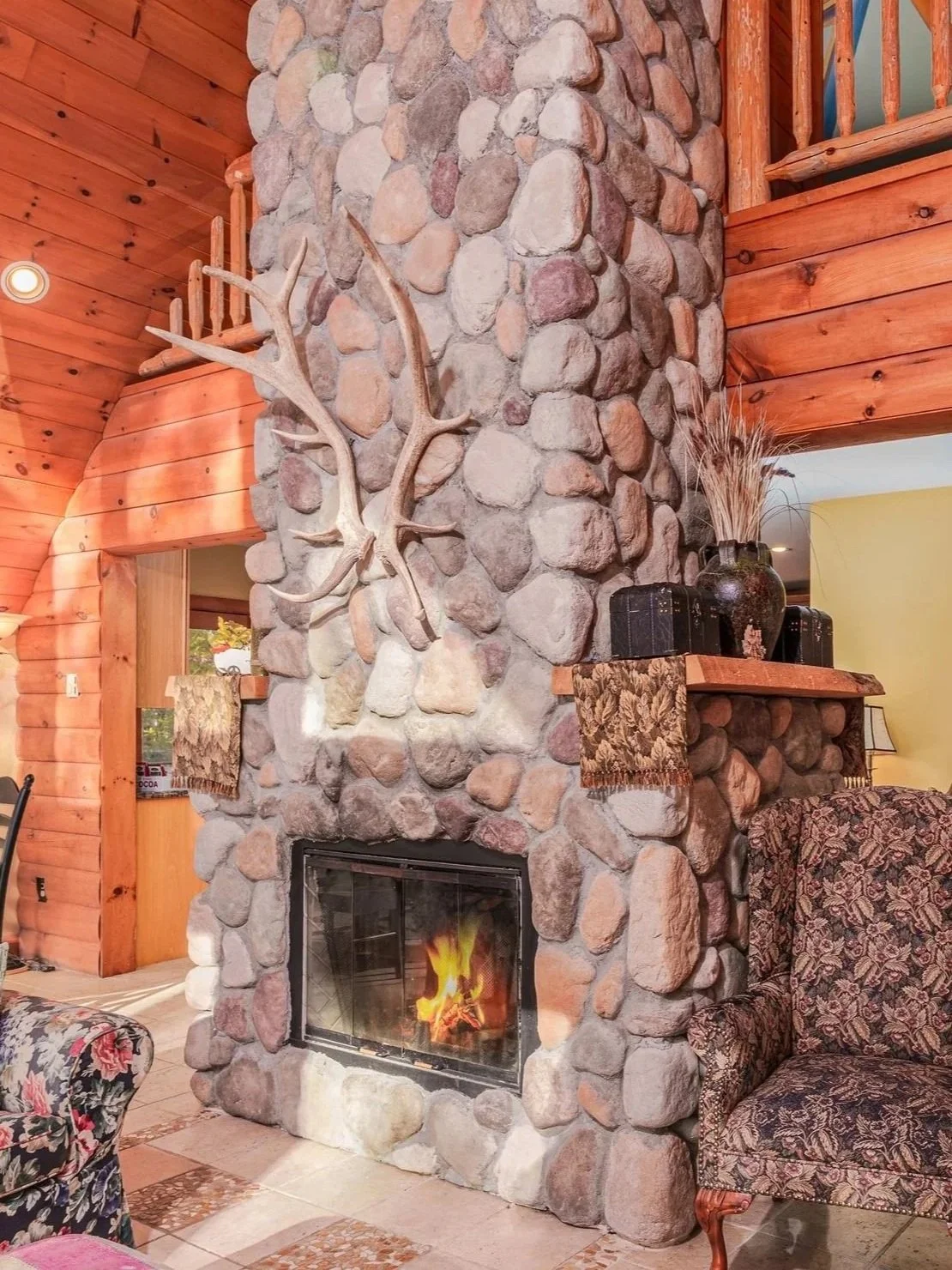 A cozy living room corner with a large stone fireplace, decorated with an antler mounted on the stone chimney, with a visible fireplace fire. The room has wood-paneled walls and a wooden mantel.