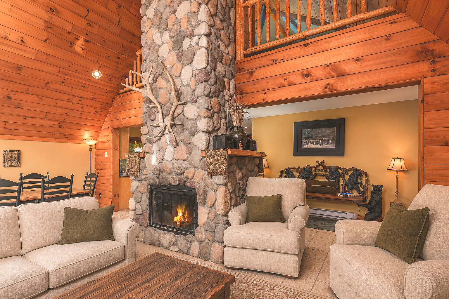 A cozy living room corner with a large stone fireplace, decorated with an antler mounted on the stone chimney, with a visible fireplace fire. The room has wood-paneled walls and a wooden mantel.