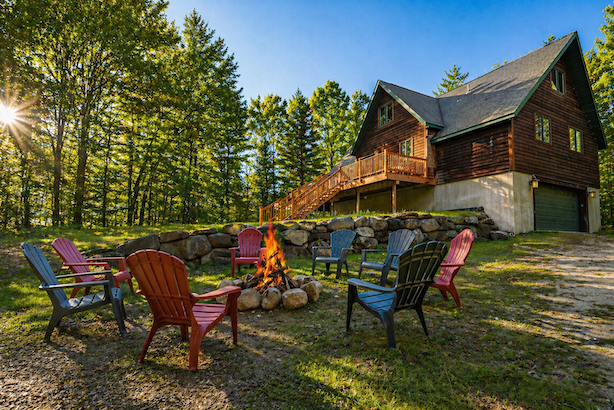 A circle of colorful Adirondack chairs surrounds a campfire on a grassy area near the 5-star lodge cabin, trees, and a clear blue sky.