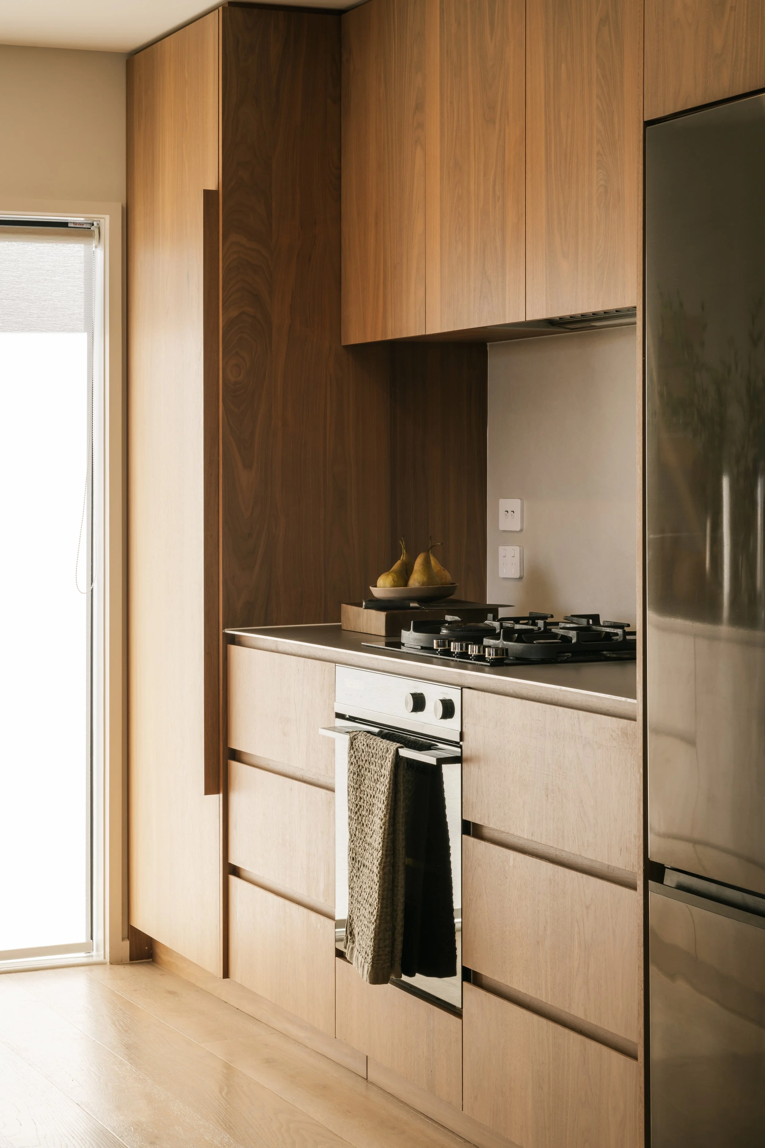 Modern Mount Maunganui kitchen with wooden cabinets, oven with a towel hanging, and a bowl with pears on the countertop near a window.
