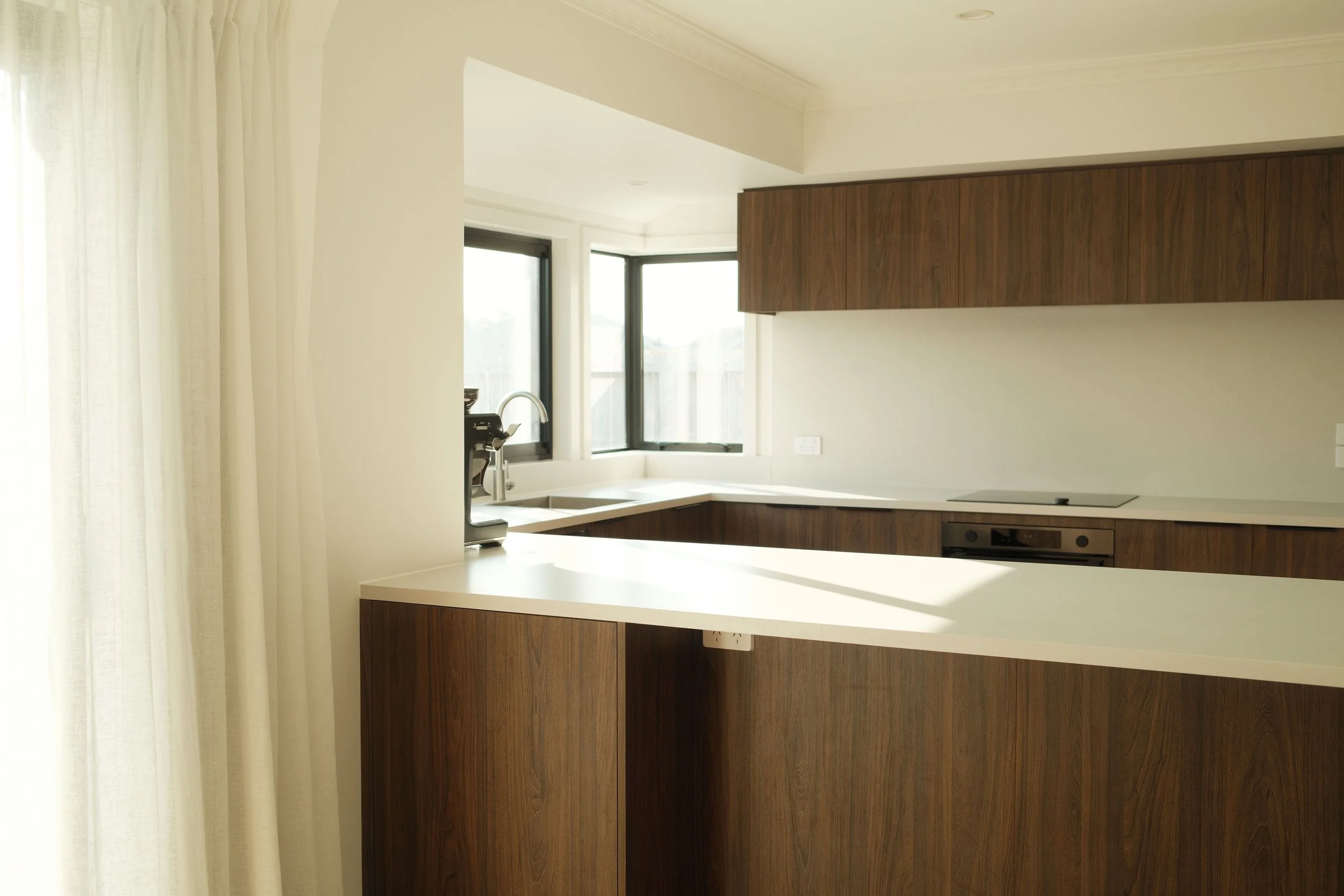 Modern Mount Maunganui kitchen with white countertops, wooden cabinets, large windows, white walls, and a black faucet near the window.