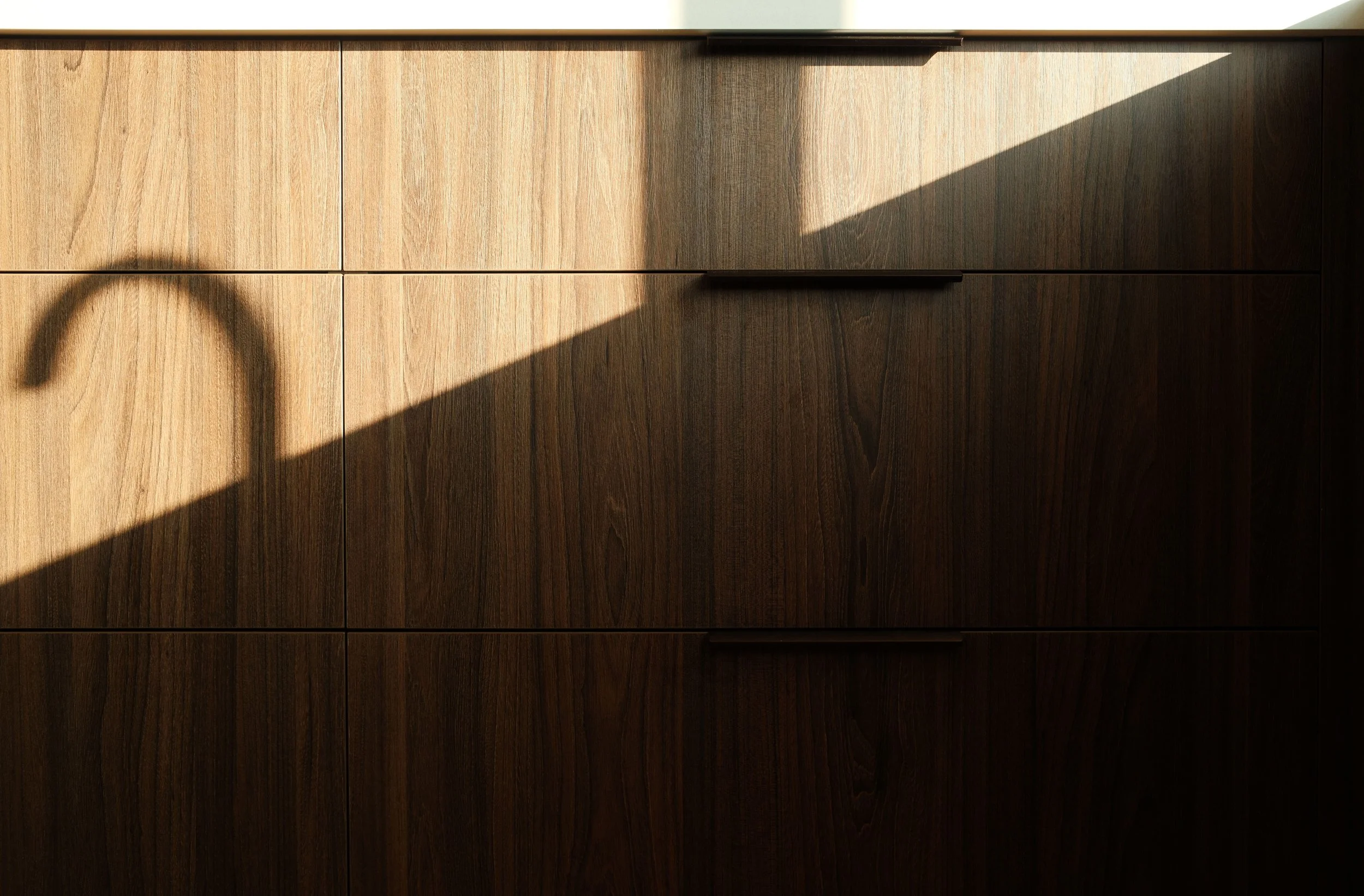 A wooden cabinet with multiple drawers and metal handles, with sunlight casting a shadow across the surface.