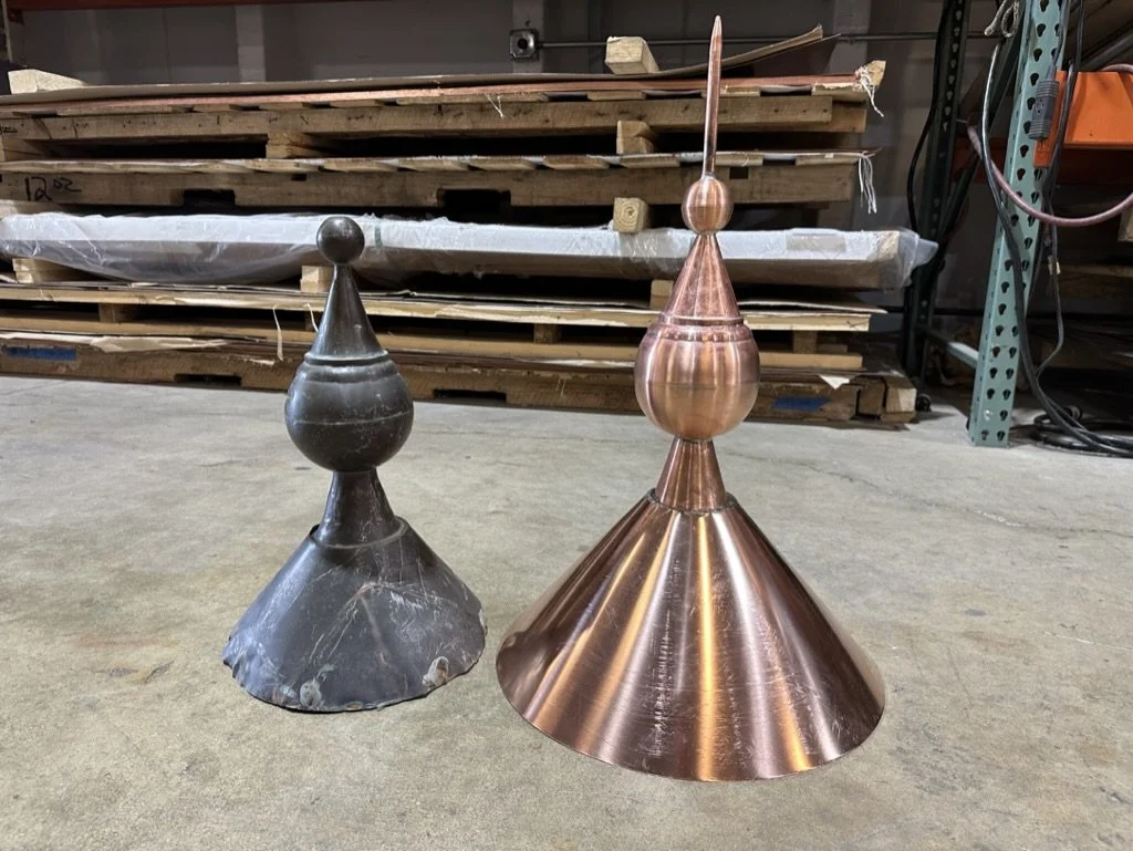 Two finials, one old / historical and one new copper reproduction, placed on a concrete floor in a workshop with wooden pallets and metal shelving in the background.