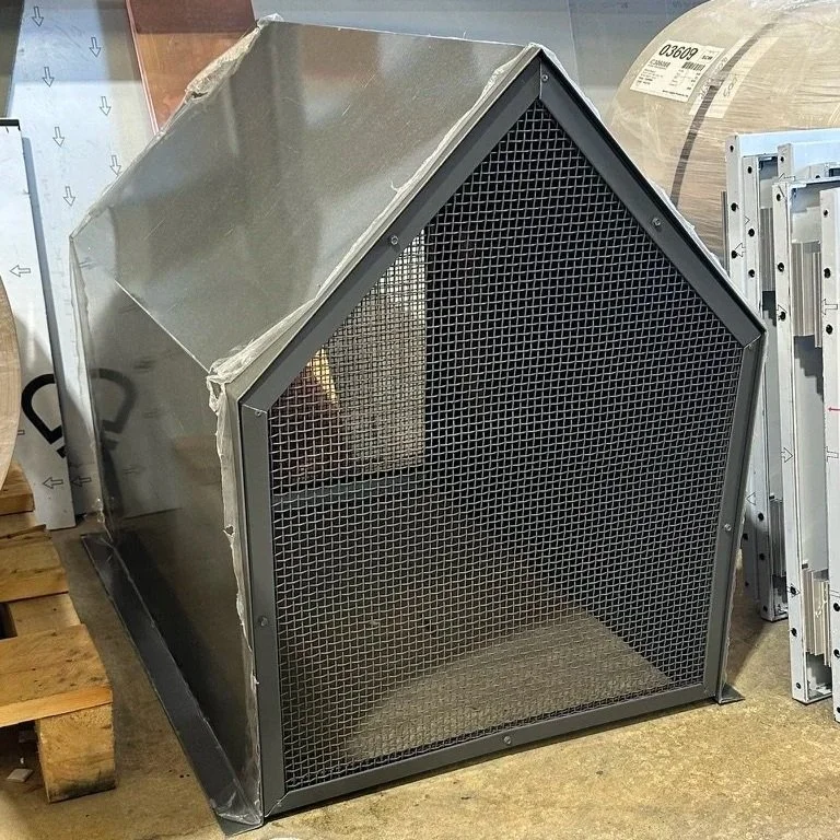 An aluminum chimney shroud with a peaked roof, mesh front, and solid sides, placed on a concrete floor.