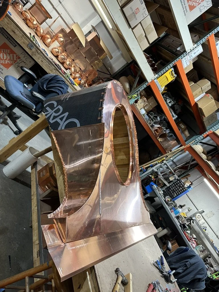 A copper dormer with a unique curved architectural design being assembled in a workshop, surrounded by tools, pipes, and storage shelves.