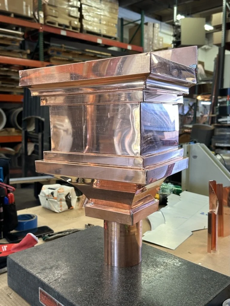 A polished copper leader head / scupper box with geometric shapes mounted on a black base, set in an industrial workshop with shelves and equipment in the background.