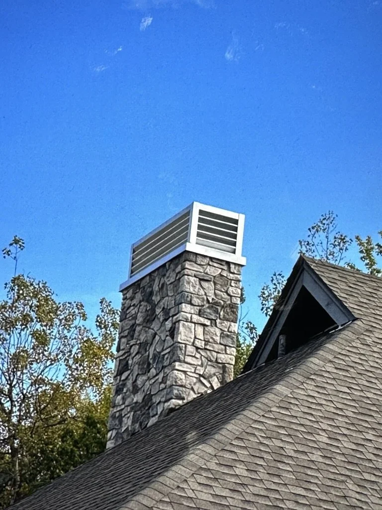 A stone chimney with a louvered shroud on top, attached to a house with a shingled roof and trees in the background, under a clear blue sky.