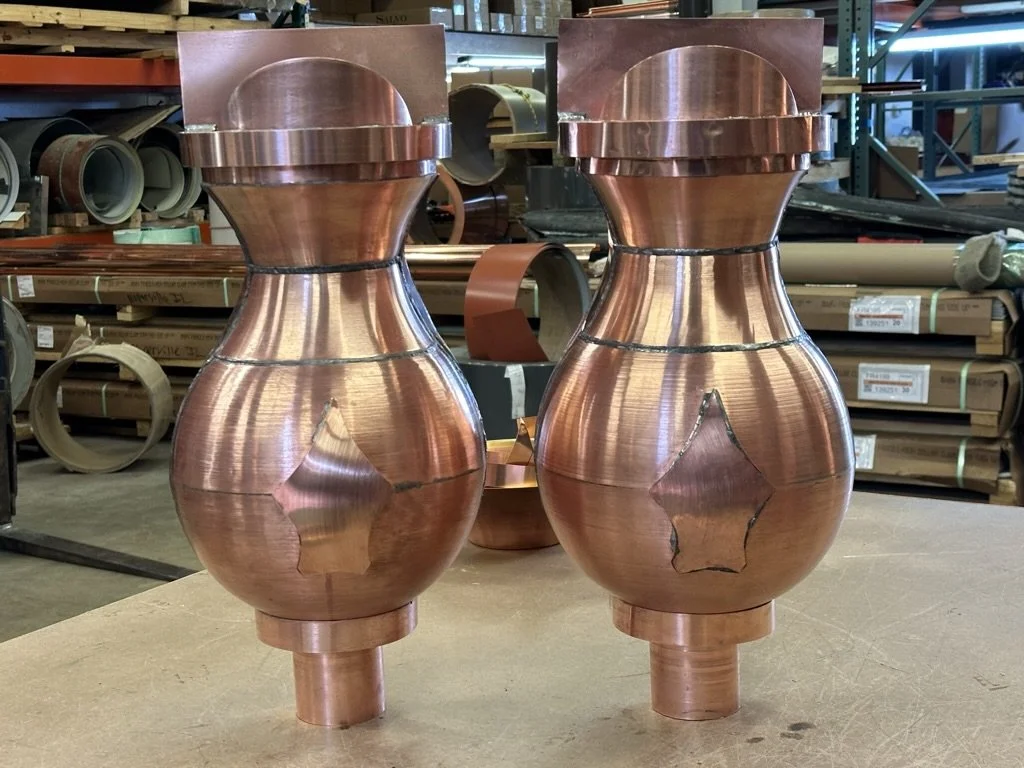 Two copper leader heads / scupper boxes, placed on a workshop table, surrounded by industrial piping and tools.