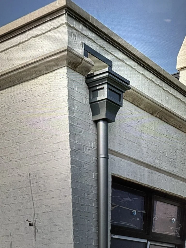 Close-up of a corner of a white brick building with a black leader head / scupper box and a rain gutter downspout.
