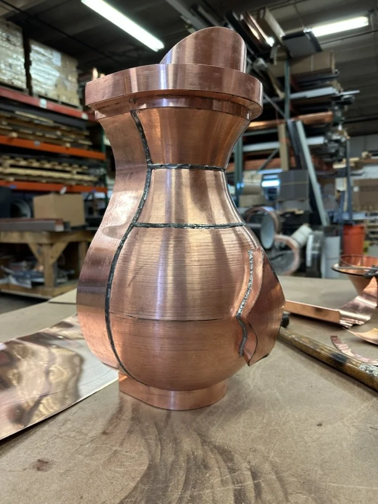 Copper leader head in a workshop, with visible solder lines and metal sheets nearby.