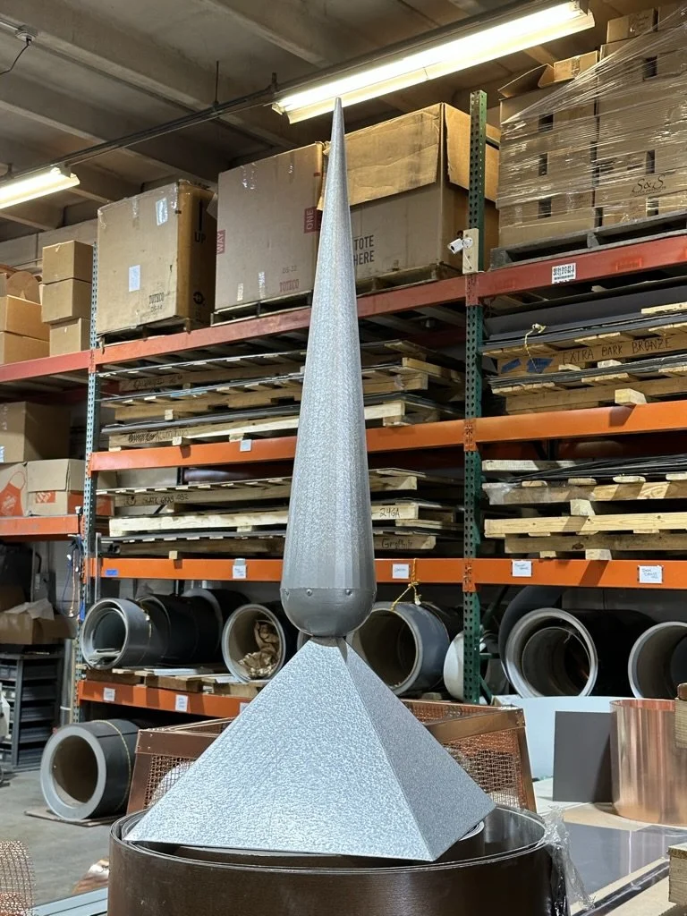 A metallic finial / spire with a tapered spiral top and a pyramid-shaped base, displayed in a warehouse among shelves with boxes and pipes.