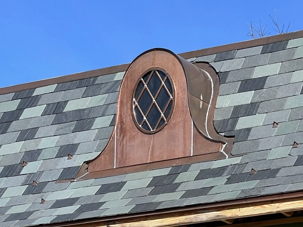 Close-up of a decorative copper dormer with a diamond-shaped grid, mounted on a gray slate roof under a clear blue sky.