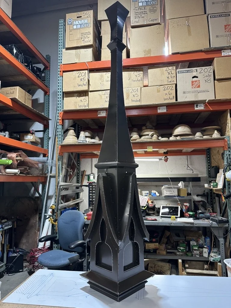 Large black metal decorative spire / finial on a workbench in a workshop, surrounded by shelving and boxes. This was a prototype of a historical reproduction project.