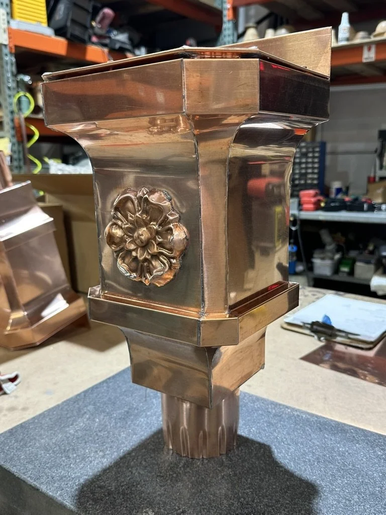 A decorative copper leader head / scupper box with floral ornamentation in the center, placed on a workbench in a workshop-like environment.