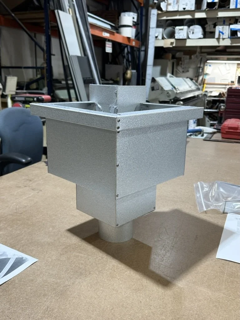 Galvalume rectangular outside corner leader head / scupper box with a rectangular inlet on a workbench in a workshop.
