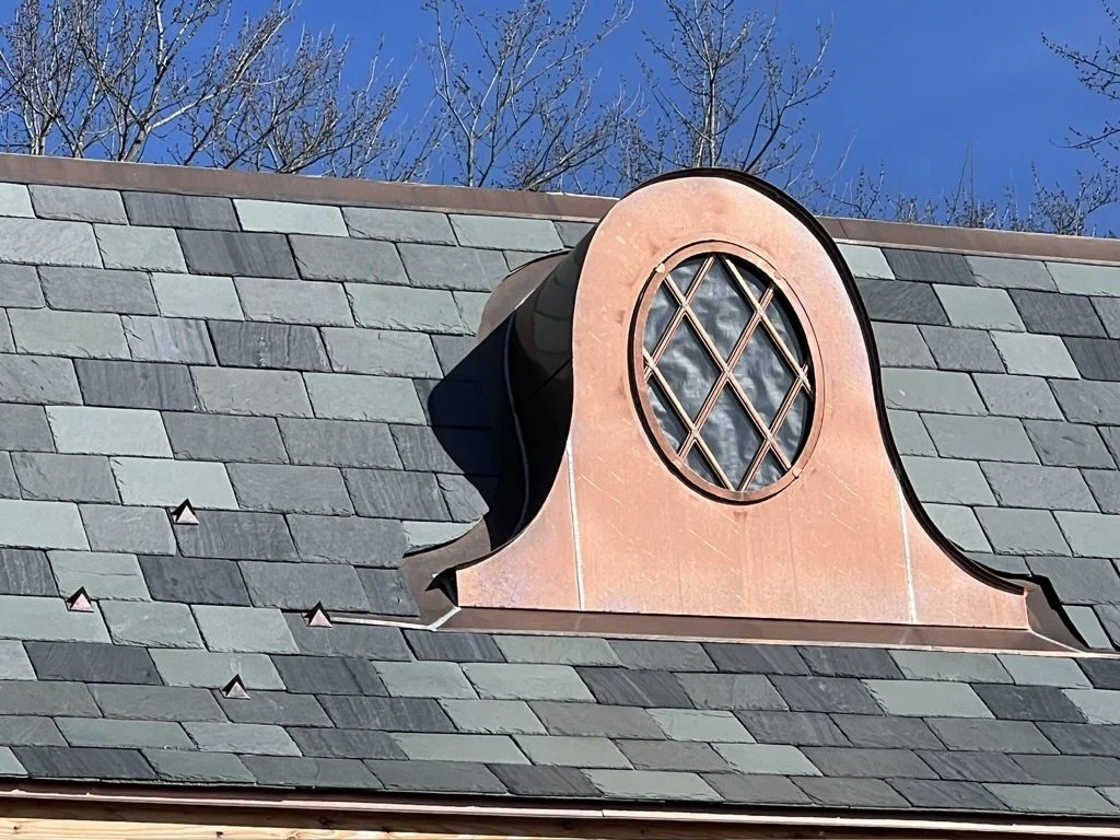 Close-up of decorative copper dormer with a circular window on a slate tiled roof, with bare trees and a blue sky in the background.