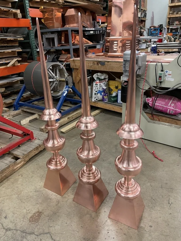 Three tall, copper finials / spires with pointed tops, ornate bases, and square bases, standing on a workshop floor.