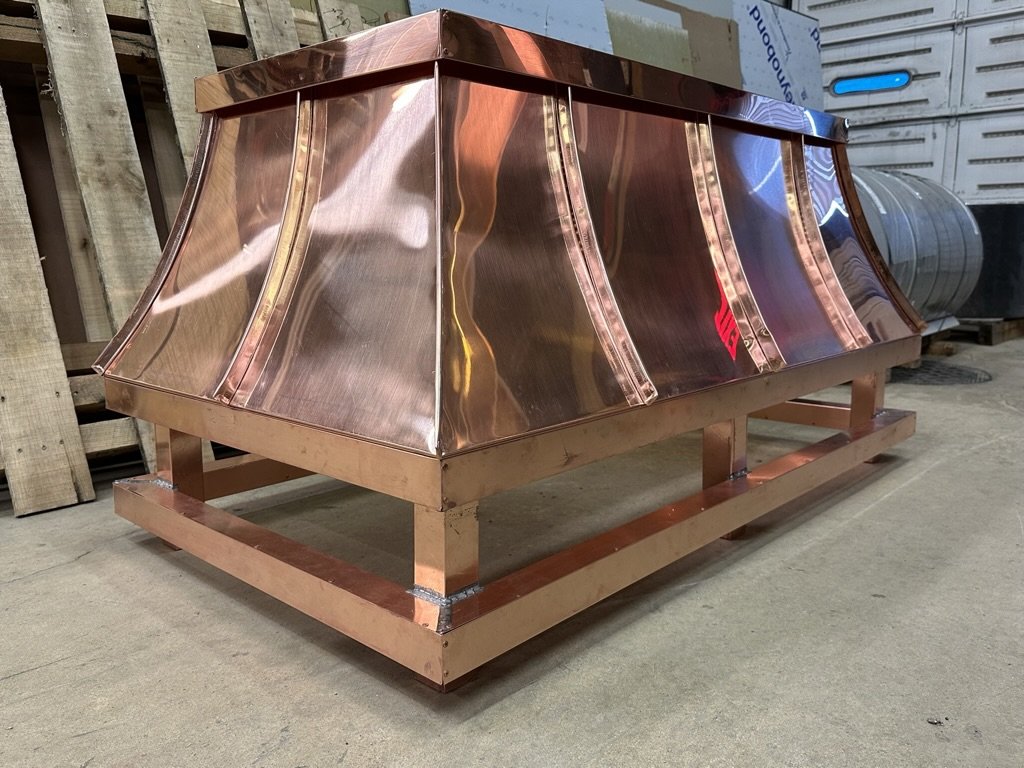 A large copper chimney shroud with a shiny, reflective surface and curved panels, placed on a wooden frame in a warehouse or workshop setting.