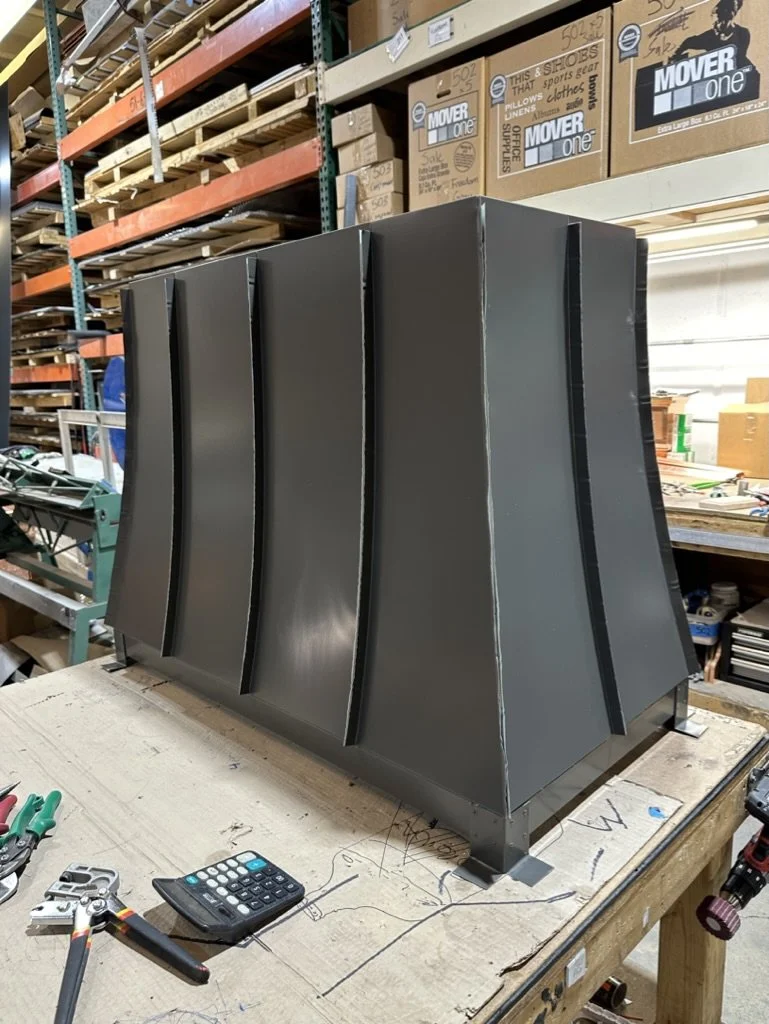 Large metal industrial style chimney shroud on a workbench in a workshop, with shelving containing boxes and equipment in the background.
